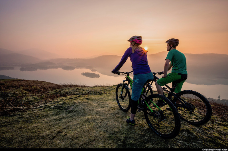 Two people on mountain bikes resting at the top of a fell above Derwentwater in Borrowdale, viewing the landscape at sunset.